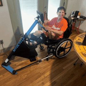 Image of a woman with an orange shirt in a black wheelchair using a bike.