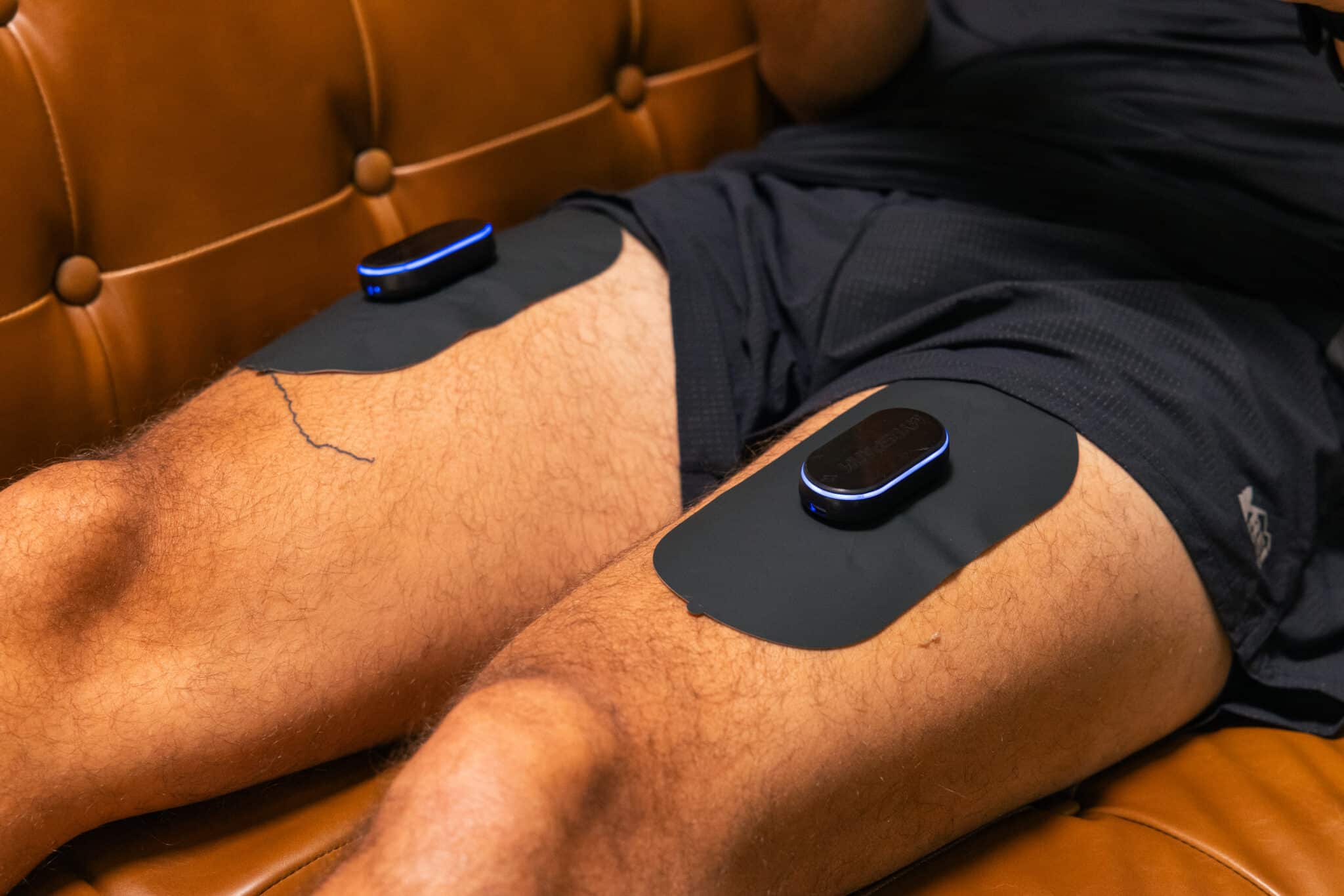 A person sits on a brown leather couch with two large black muscle stimulation pads, each featuring a small electronic device on top—demonstrating one of the types of electrical stimulation used for muscle therapy.