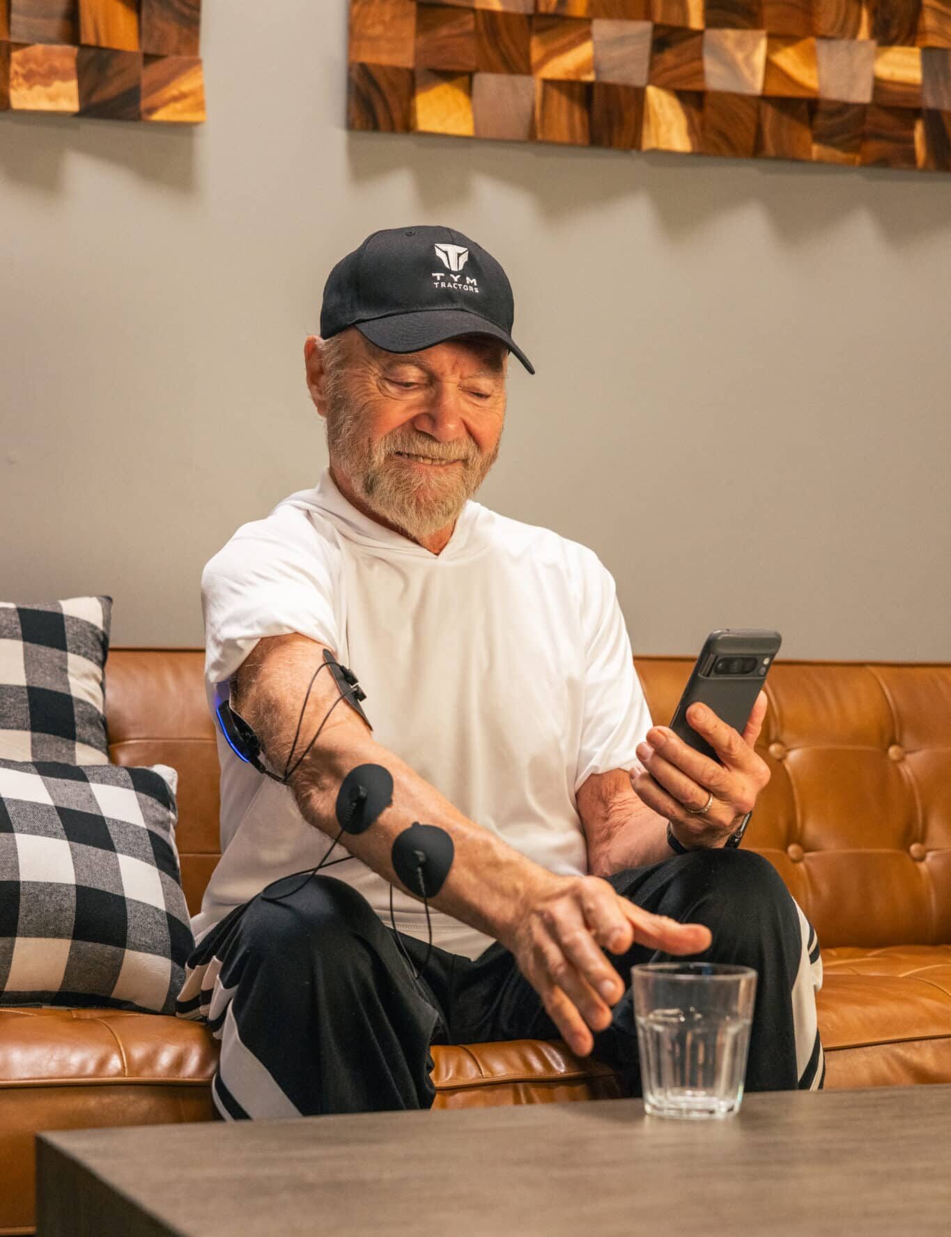 An older man sits on a brown leather couch using a smartphone. Electrodes are attached to his arm, and he reaches for a glass of water on the table in front of him, smiling.