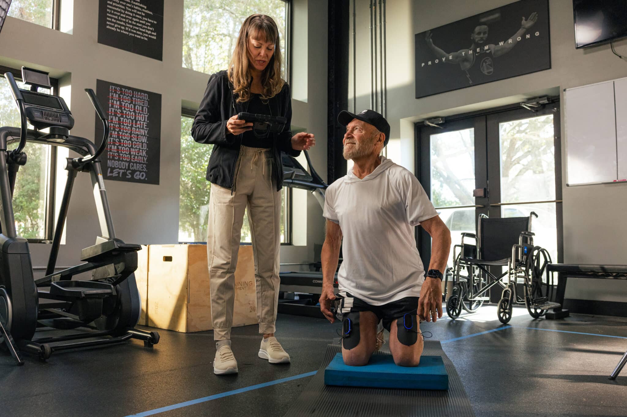 A man with prosthetic legs, including a MyoSpark foot drop device, kneels on a blue mat in a gym while a woman stands next to him holding a tablet. Fitness equipment and a wheelchair are visible in the background as they engage in conversation.