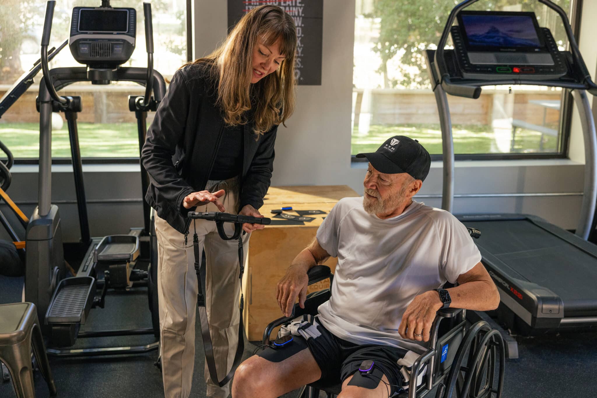 A woman stands next to a man in a wheelchair at a gym, showing him something on a tablet. They appear engaged and smiling, discussing types of paralysis as exercise equipment and treadmills are visible in the background.