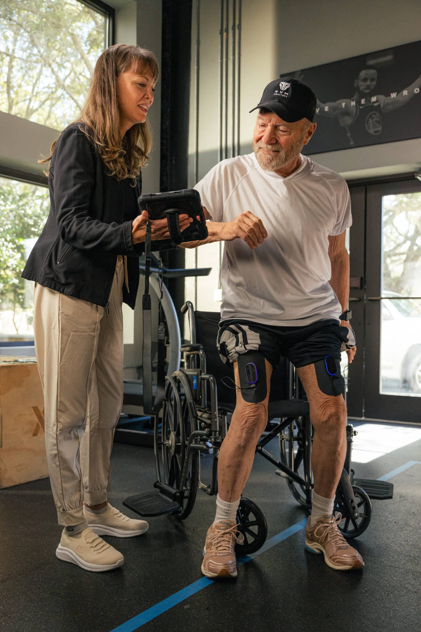 A smiling woman shows a tablet to an older man standing beside a wheelchair, wearing knee braces and a cap, inside a bright room with large windows.