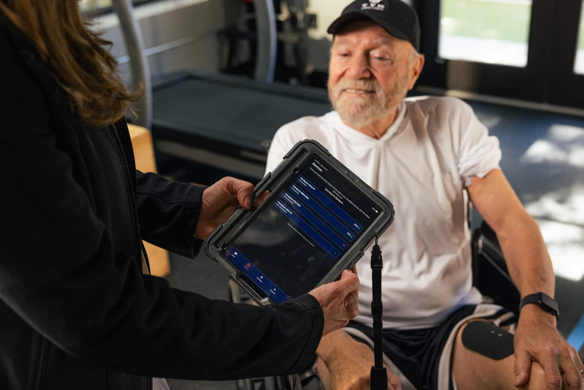 A woman holds a tablet displaying a digital interface in front of an older man in sportswear and a cap, seated in a wheelchair, as EMS muscle stimulator equipment is visible in the background.