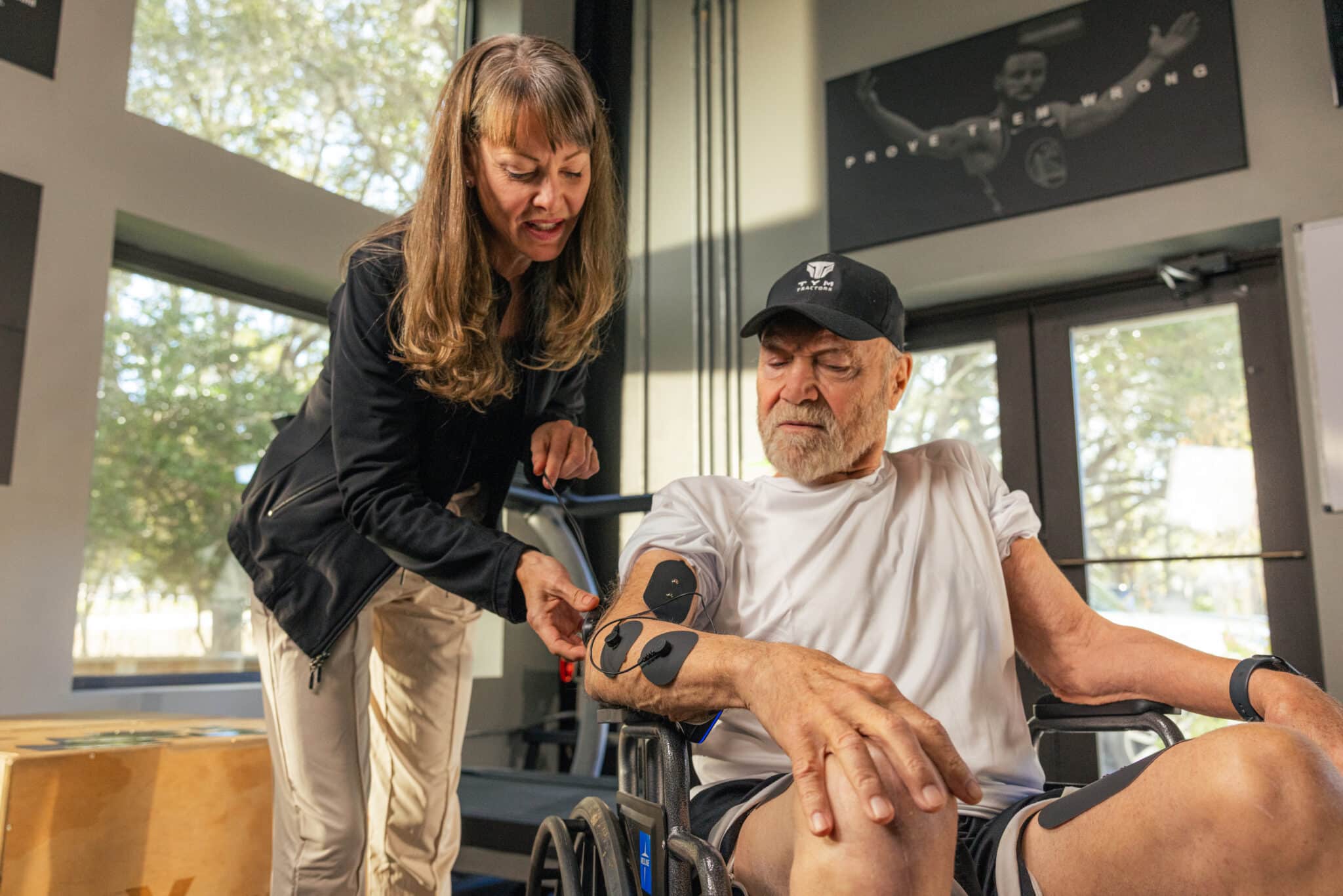 A woman demonstrates MyoSpark Fit use cases by assisting an older man in a wheelchair, adjusting electronic muscle stimulation pads on his arm in a bright, modern gym. Both appear focused on the process.