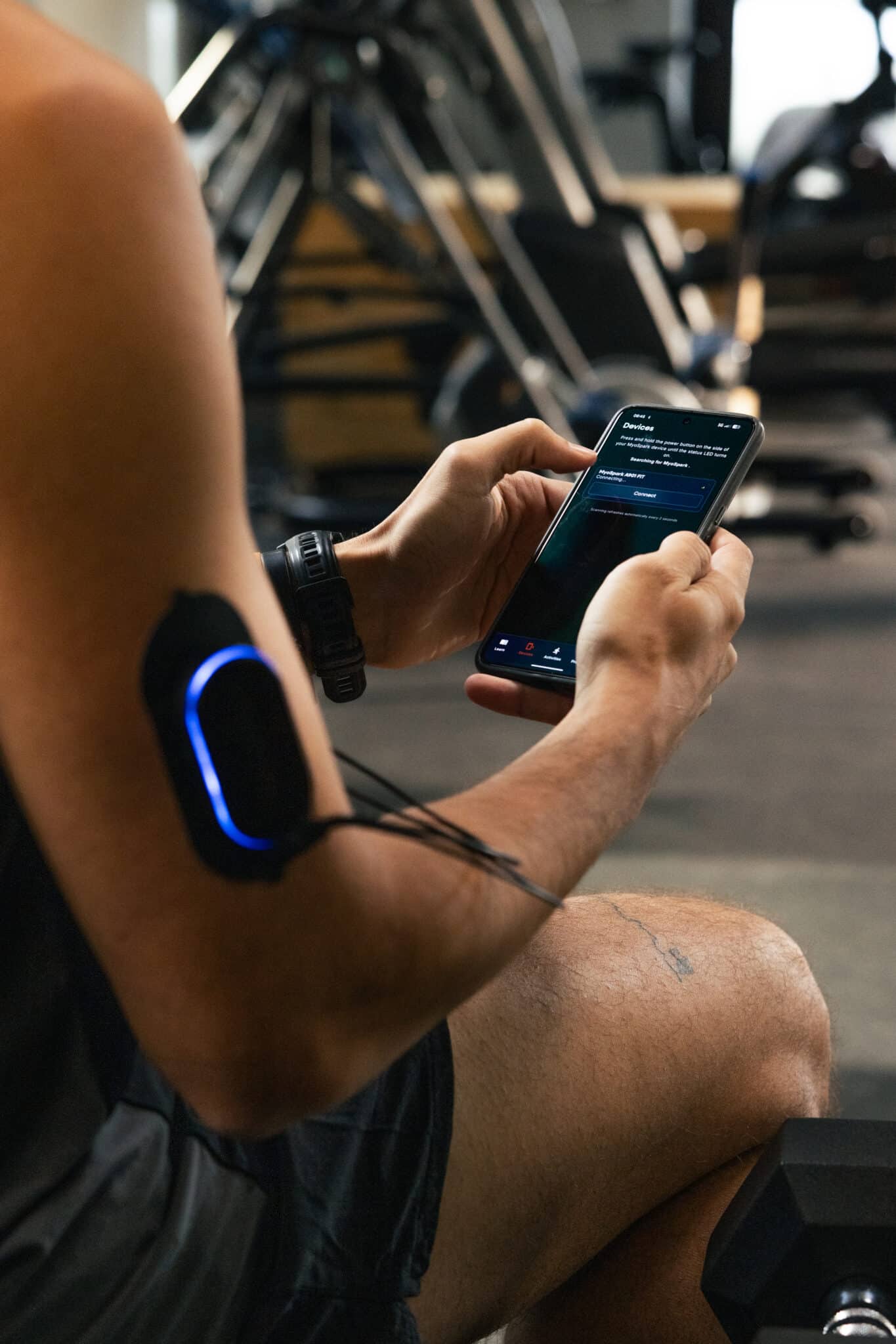 A person in workout attire sits in a gym, using a smartphone. An electronic muscle stimulation device with glowing blue light is attached to their upper arm, with wires connected—nearby, gym equipment and an FES bike are visible in the background.