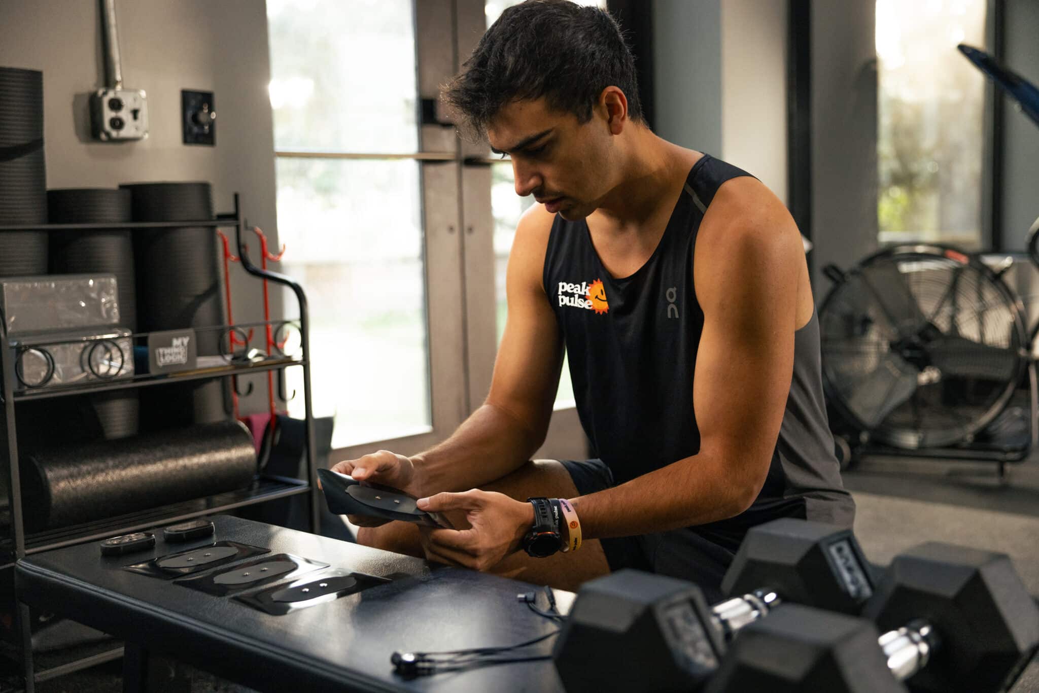 A man in athletic wear sits at a table in a gym, assembling or examining fitness equipment parts—demonstrating practical MyoSpark Fit use cases. Dumbbells, exercise mats, and a large fan are visible in the background.