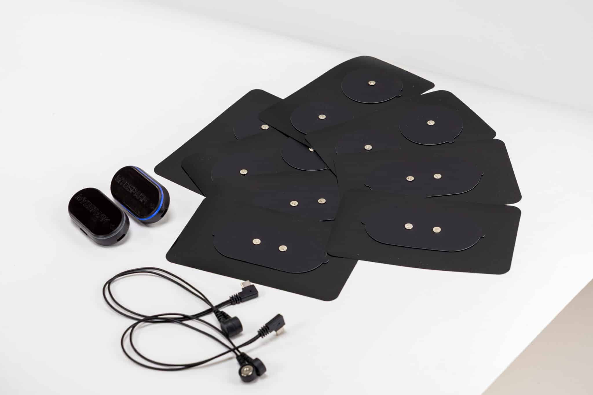 A set of eight rectangular black electrode pads, two small Fit Fitness electronic devices, and a black cable with connectors are arranged on a white surface.