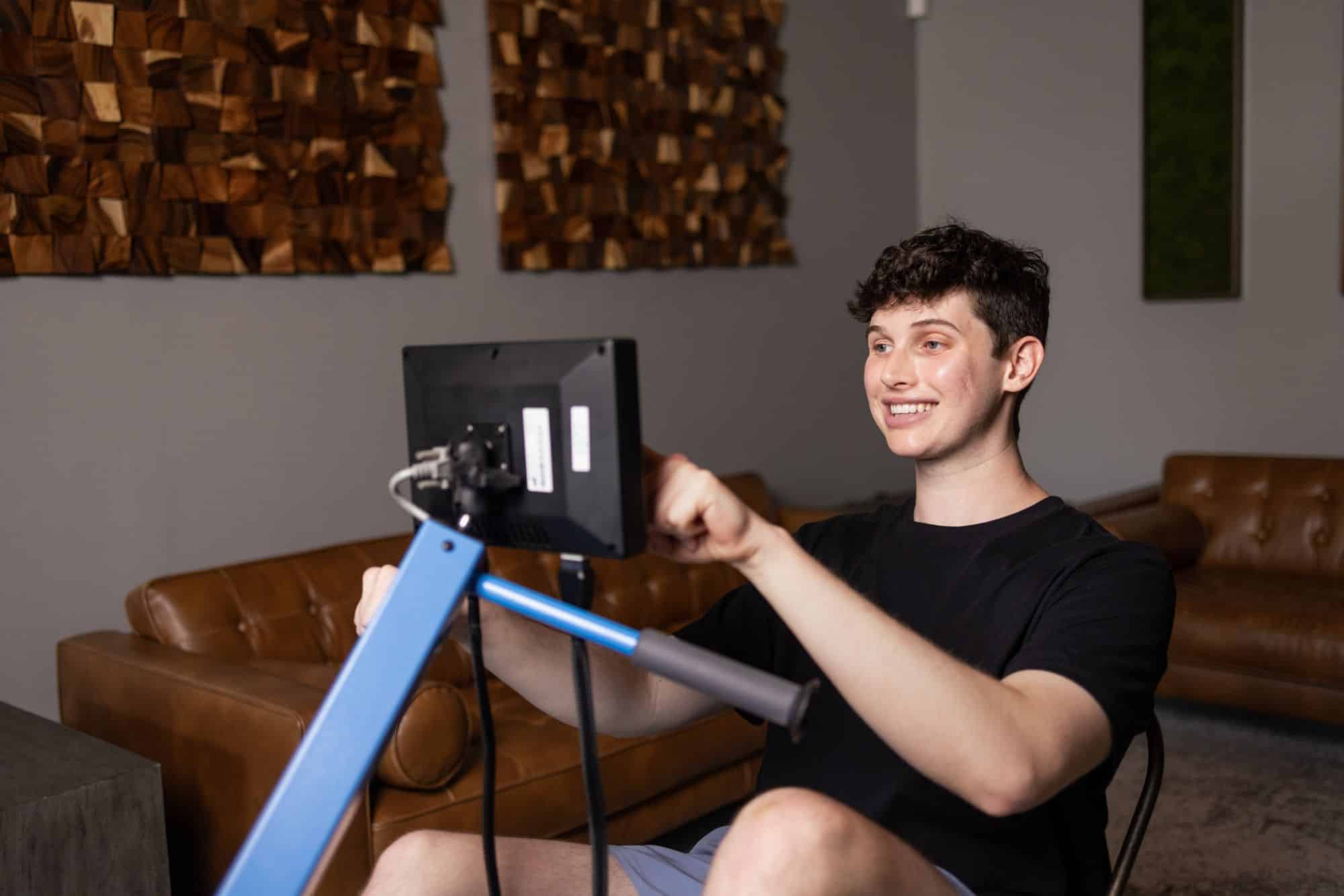 A young man sits indoors, smiling and using a MyoCycle FES bike with a digital screen. He wears a black t-shirt and shorts, while brown couches and wooden wall panels are visible in the background.
