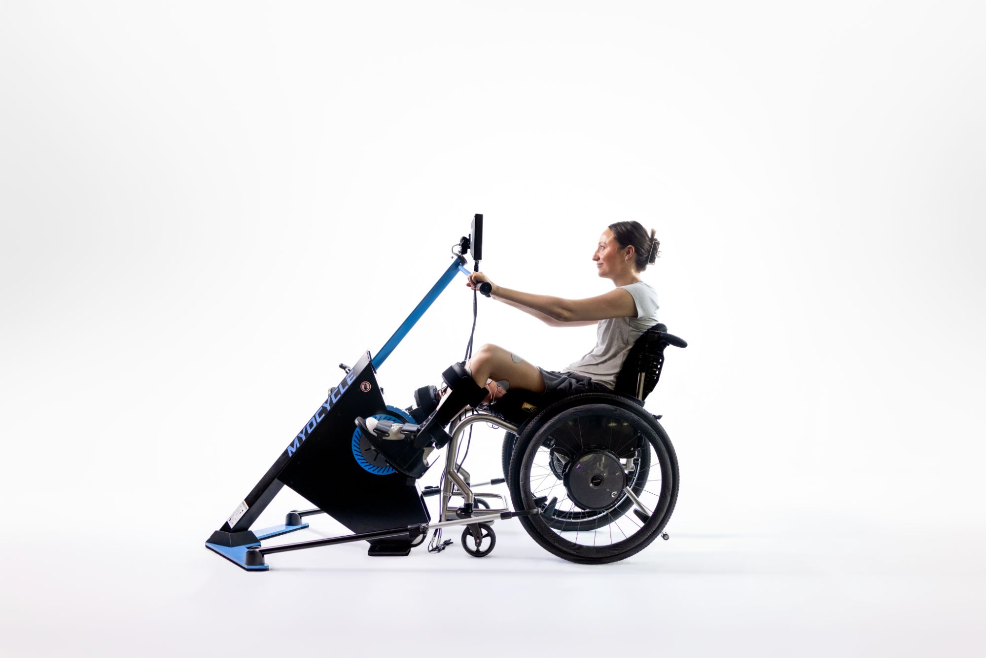 A woman in athletic clothing sits in a wheelchair, using a stationary handcycle exercise machine for FES cycling research against a plain white background.