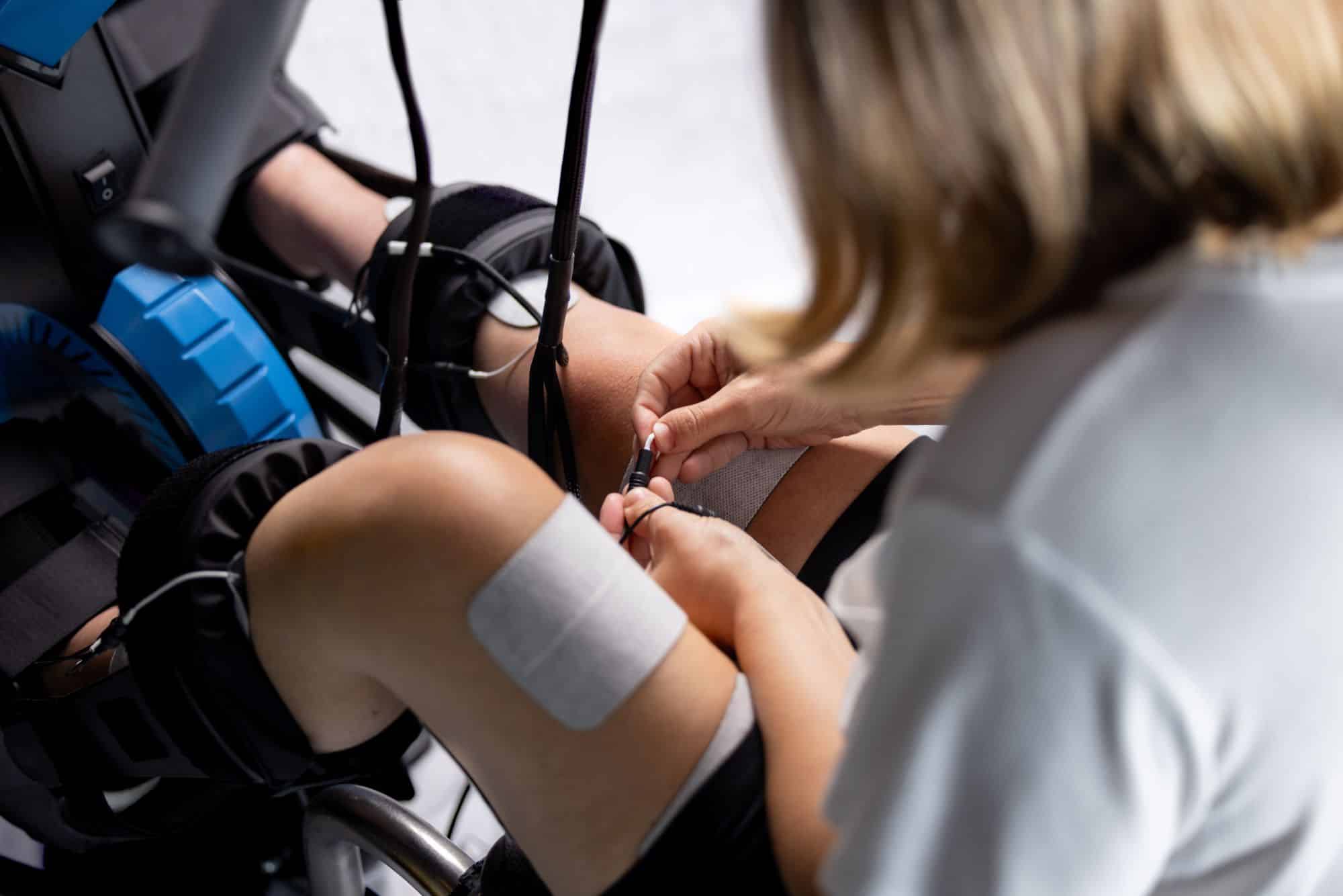 A person applies electrode pads to another person's knees, possibly using one of the types of electrical stimulation for physical therapy or rehabilitation, with supportive equipment visible in the background.