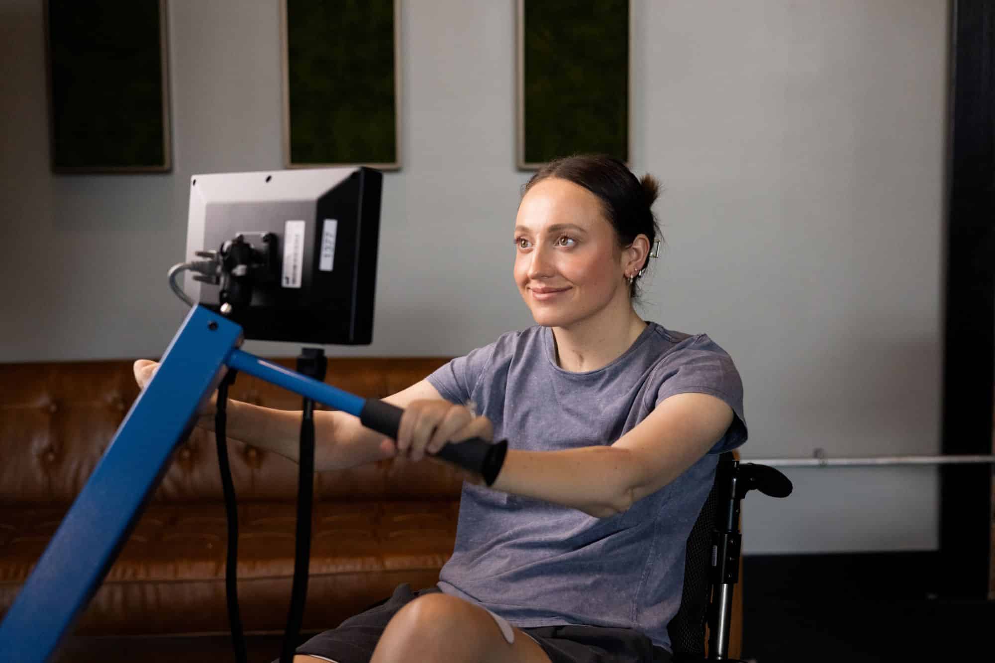 A woman in a wheelchair exercises on a rowing machine indoors, looking at a screen in front of her and smiling.