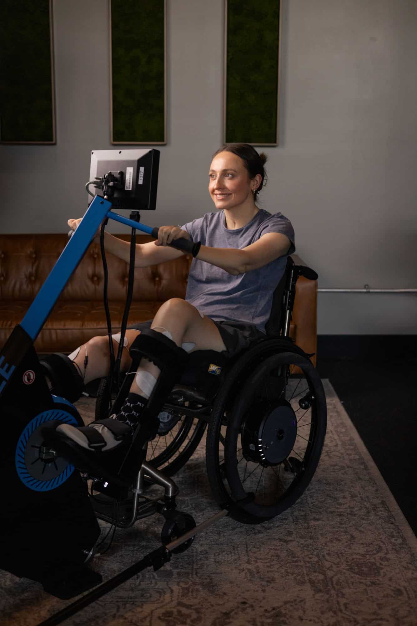 A woman in a wheelchair, wearing braces on her legs, uses an FES bike with a screen in a comfortable indoor setting. She is smiling and engaged in the activity.