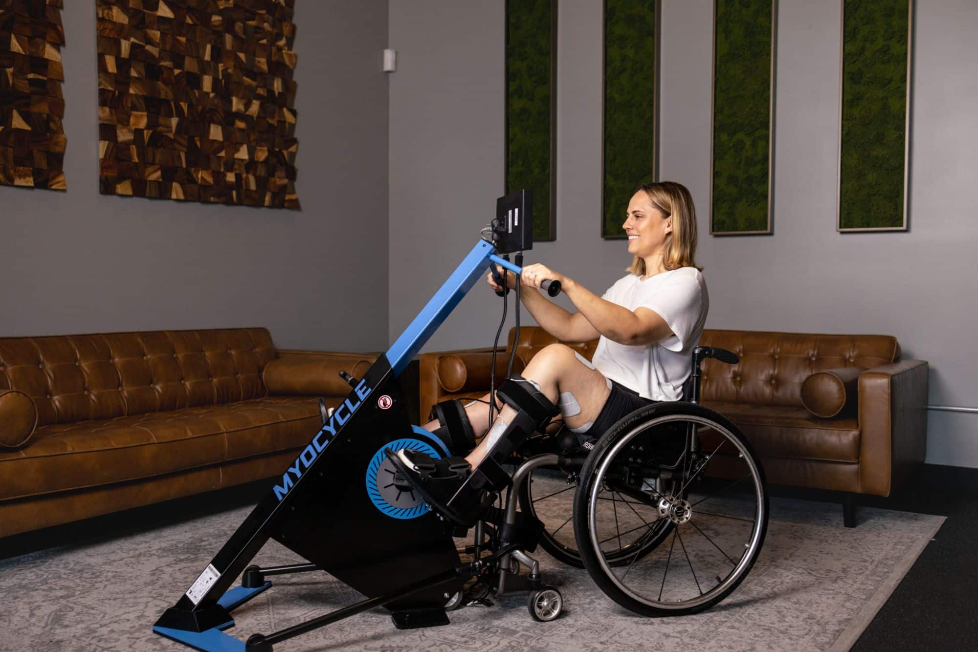 A woman in a wheelchair uses a MyoCycle exercise machine in a modern room with brown couches, a gray rug, and green wall panels. She is holding the machine’s handles and has her feet strapped to the foot pedals.