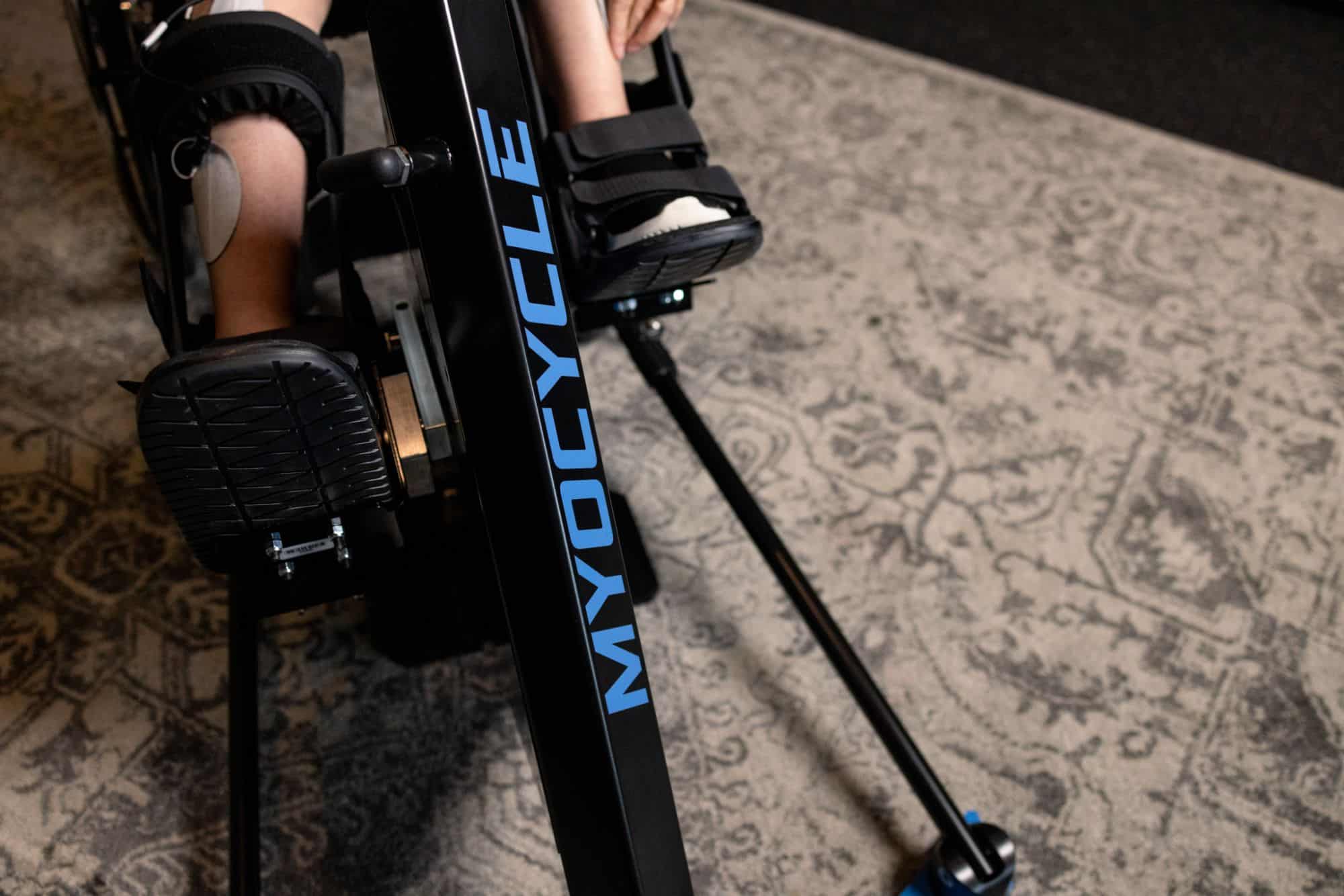 Close-up of a person’s legs and feet strapped into a black stationary exercise bike labeled “MYOCYCLE” on a patterned rug.