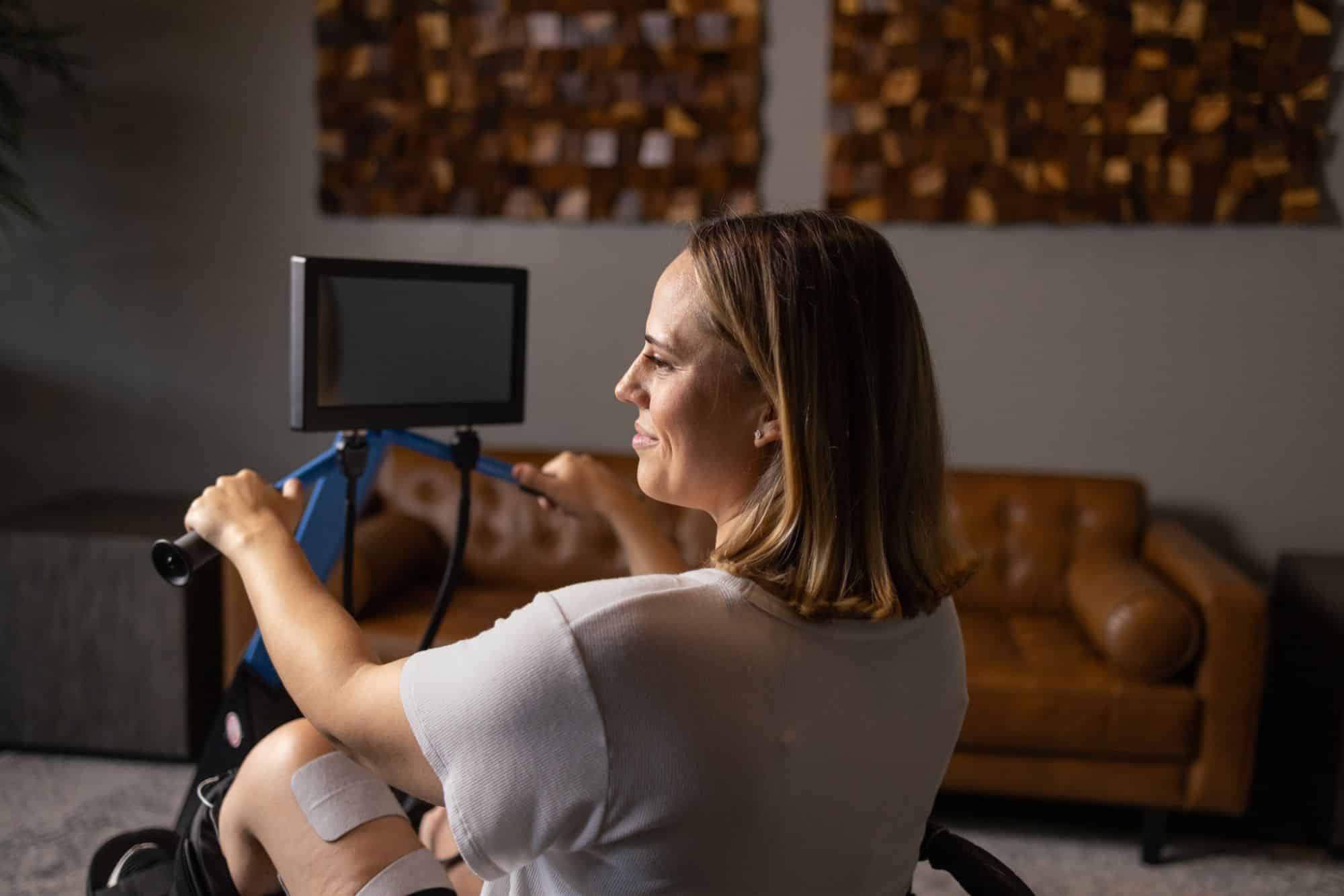 A woman sits and exercises on the MyoCycle Pro, smiling and looking forward, with a brown couch and abstract wall art in the background.