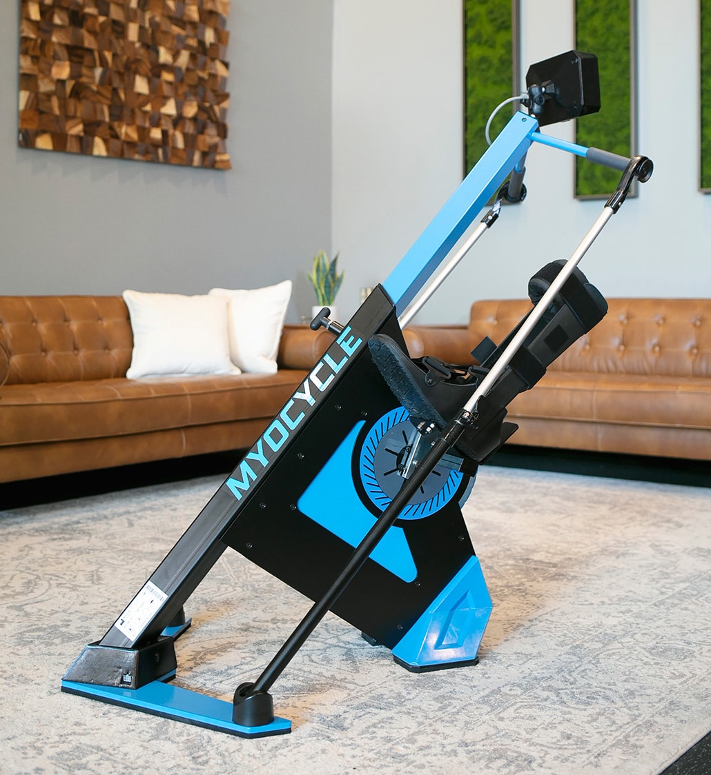 A blue and black MyoCycle FES bike sits on a rug in a modern living room with a brown tufted sofa, white pillows, wall art, and green decor in the background.