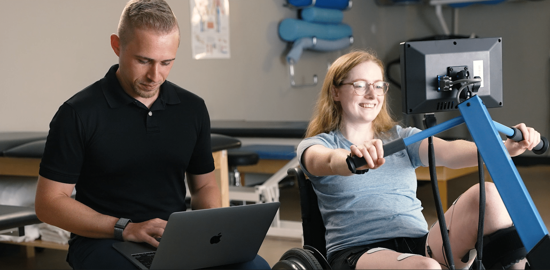 A young woman in athletic wear uses a MyoCycle Pro handcycle machine while smiling. A man beside her, wearing a black polo shirt, types on a laptop. Therapy and exercise equipment are visible in the background.