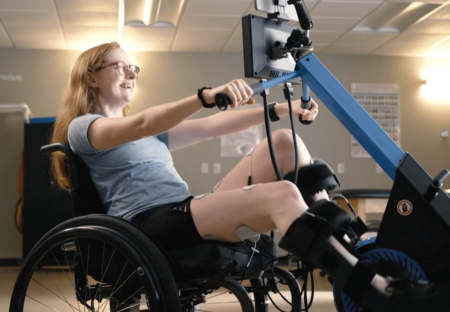 A woman in a wheelchair uses the MyoCycle Pro rowing machine in a gym. She is smiling and wearing exercise clothes, with adaptive equipment on her legs. The room features gym equipment and overhead lighting.