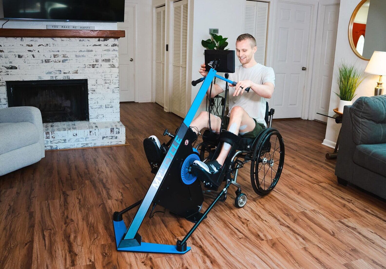 A man in a wheelchair exercises with the MyoCycle Home stationary handcycle machine in a living room with wooden floors, a couch, a lamp, a fireplace, and plants.